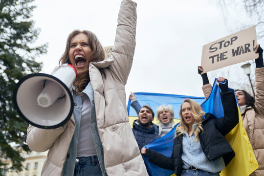 Caucasian woman screaming through megaphone in front and group of young people manifesting against Ukrainian war in the background