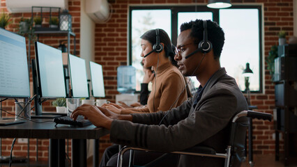 Employee with impairment working at customer care service to give support to people needing help. Man in wheelchair sitting at call center workstation to talk to clients about telemarketing.