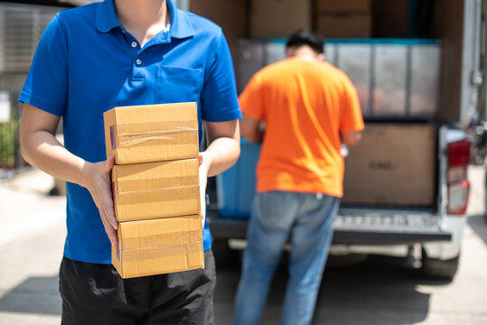 Asian Delivery Man Wearing Face Mask And Glove In Blue Uniform Holding A Cardboard Boxes In Front House, Copy Space. Truck Movers Loading Van Carrying Boxes And Moving House. Courier Wearing Mask.