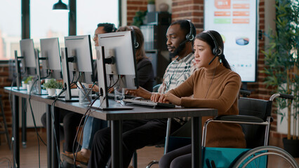 Asian woman with disability using headset to help clients at call center, talking to people about telemarketing. Employee in wheelchair working at customer assistance service. Tripod shot.