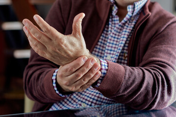 A man sitting at home is holding his wrist because he is in pain due to osteoarthritis, arthritis,...
