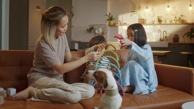 Young Asian Mother and Little Siblings Spend Time with Dog at Modern Flat Play Together Seated on Sofa, Enjoy Playtime Use Rainbow Wooden Toys. Babysitting Work, Leisure at Home with Kids Concept