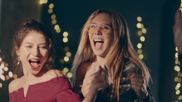 Close up couple of young multiracial girls wearing plush dresses enjoying glamorous party and having fun. Women dancing with falling confetti and sparkling or glowing sparklers or fireworks.