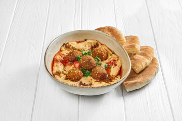 Hummus with vegetables and falafel in a plate on a white wooden background.