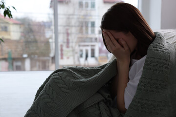 Fototapeta premium Sad young woman wrapped in warm plaid crying at home, space for text. Loneliness concept