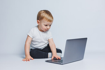 A little boy 3-4 years old sits with a laptop on a white background. A child in a white T-shirt and black trousers looks at a laptop. Modern children