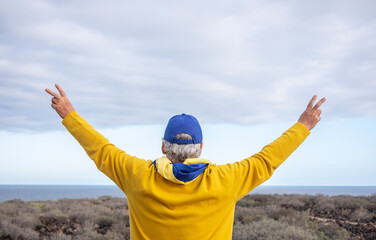Rear view of senior man standing in front of the sea with the Ukrainian flag around his neck with...
