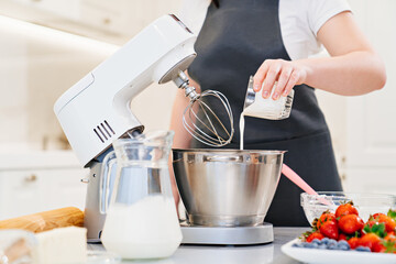 A women cook adds a milk to prepare dough or cream in a mixer bowl.