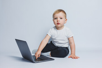 A smart boy 3-4 years old sits with a laptop on a white background. A child in a white T-shirt and black trousers sits at a laptop and looks at the camera. Modern children