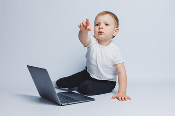 Little smart boy 3-4 years old sits with a laptop on a white background. A child in a white T-shirt and black trousers sits at a laptop and looks at the screen. Modern progressive children