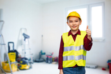 building, construction and profession concept - little boy in protective helmet and safety vest showing thumbs up over room with working equipment background