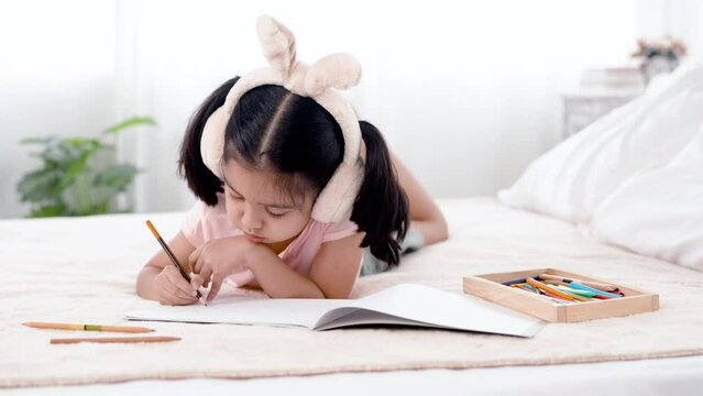 4K 50fps, adorable Asian girl tied her hair and wearing bunny ears, happily laying on a white bed drawing pictures..