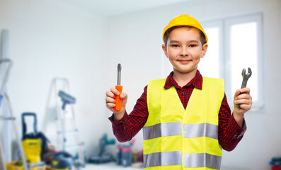 building, construction and profession concept - little boy in protective helmet and safety vest holding screwdriver and wrench over room with working equipment background