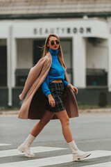 Blonde in fashionable clothes with skirt and blue sweater walks down the city street in sunglasses. Selective focus