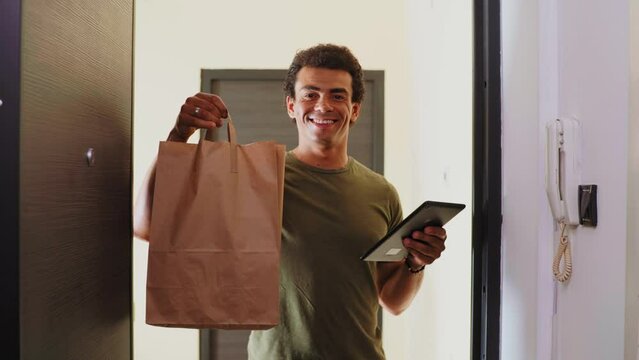 Smiling delivery man in front of the door with tablet and paper bag