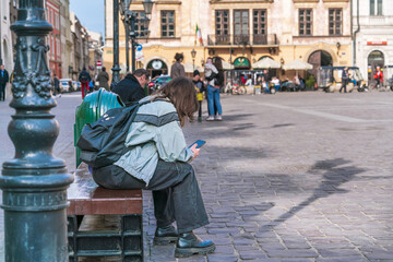 Fototapeta premium A young girl is sitting on a bench in the city square with a backpack on her back and a smartphone in her hands on a sunny day
