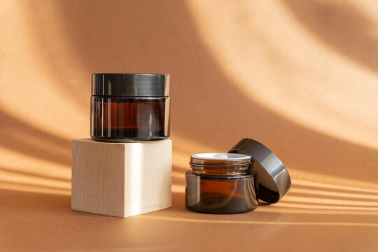 Two jars of amber glass for cosmetics on a brown background with shadows in the form of stripes. Mock-ups of containers with moisturizer for face, hands, on wooden square 3d stand