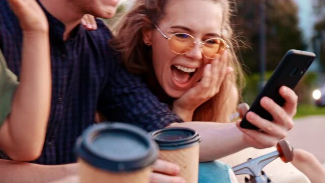 Happy Smiling Teenage Friends Laughing Outside At Something In Smartphone Or Mobile Phone. Three Young Multiracial People Spending Time Together. Friendship, Communication, Youth And Lifestyle Concept