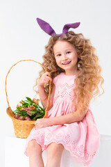 Happy holiday easter kids. cute little beautiful girl with rabbit hare bunny ears, basket of chocolate eggs, spring flowers on white background, vertical