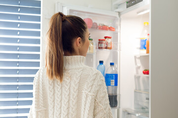 Young woman in a sweater peeks into the refrigerator