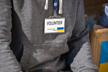 Cropped photo of volunteer young man with badge for helping ukranian people refugees sitting near the boxes with the food.