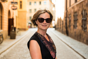 Obraz premium Portrait of a smiling thirty year old white woman with a black dress and sunglass in the Sunny Streets of Stockholm Old Town