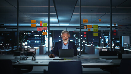 Successful Senior Businessman Working on Laptop Computer in Big City Office Late in the Evening. Investment Analyst Checking Financial Graphs from Project Management Reports.