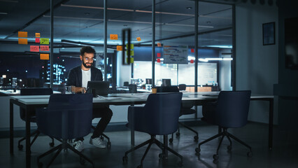 Successful Handsome Creative Director Working on Laptop Computer in Big City Office Late in the Evening. Businessman Preparing for a Marketing Plan Meeting Discussion  in Conference Room.