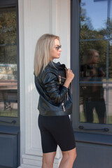 walk activity. blonde girl in black leather jacket and cycling shorts is standing in profile near park cafe background and looking away with smile. sport lifestyle concept, free space