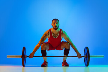 Studio shot of young man in red sportswear exercising with barbell isolated blue background in neon. Sport, weightlifting, power, achievements concept