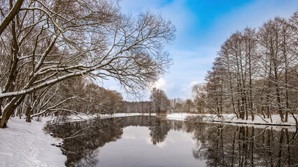 Winter landscape. The river surface of the water, blue sky, snow on the banks, reflections of trees without foliage in the water.