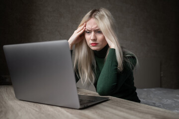 A sad and very tired blonde woman in a green golf is bored working at home with a laptop. Girl sitting at the table in a bad mood in the room. Online learning. Unsightly video conference. High quality