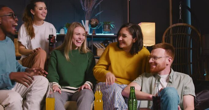 Group of happy fun multiethnic young friends talk, laugh cheerfully sitting on couch together with drinks at cozy home.