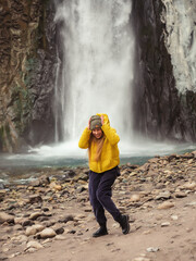 A woman stands crouched and laughing on the river bank against the backdrop of a stormy waterfall...