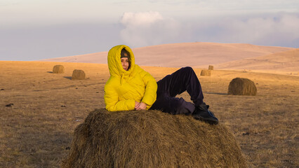 A woman in a warm yellow jacket lies on a haystack in a field. Caucasian countryside