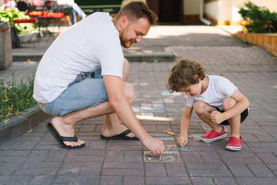A little preschool boy with father draws with colorful chalks on the ground. Positive happy toddler child drawing and creating pictures. Creative outdoors activity in summer.