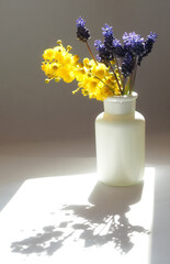 White ceramic vase with yellow and blue flowers on the table. Sunlight and shadows
