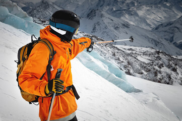 A professional skier athlete in a red jacket, helmet and mask stands on a slope against the...