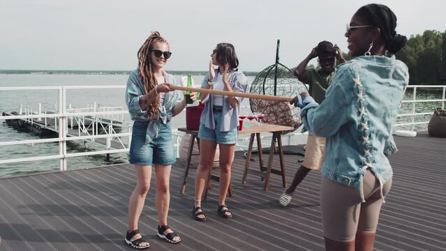 Young friends having party on lake pier, playing limbo, drinking beer, laughing and clapping hands on hot summer day