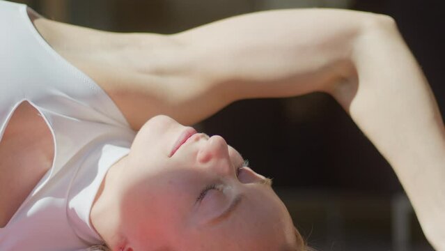 Long hair blond white young woman is practicing yoga or pilates in a white t-shirt doing inverse position with her arm above the head. Strong hand, biceps. Sun light falls on her. Close-up video 