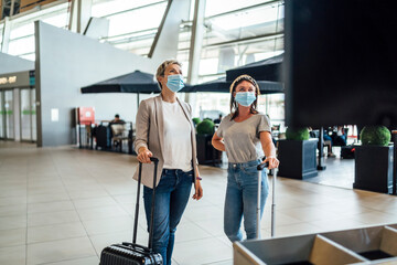 Two travelling women wearing protective masks discussing by flight information board at the airport