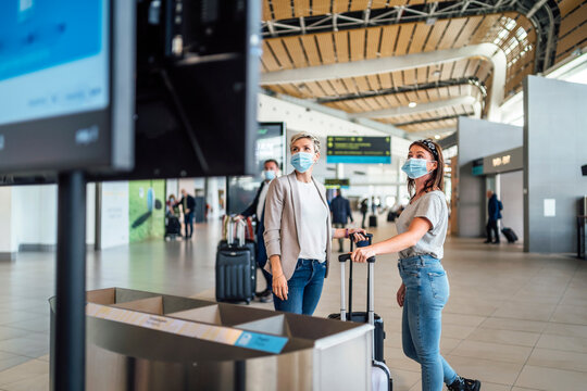 Two travelling women wearing protective masks discussing by flight information board at the airport