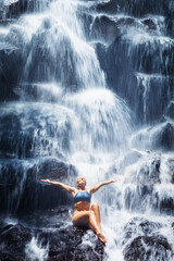 Fototapeta na wymiar Travel in Bali jungle. Beautiful young woman sitting on rock under falling spring water, enjoy tropic cascade waterfall. Asian nature, day trip, walking adventure, fun on family summer vacation
