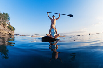 Young happy couple have fun on stand up paddleboard. Active paddle boarder paddling by sunset sea....