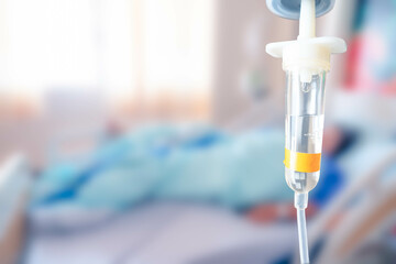 Close up of saline intravenous (IV) drip with blurred patient on bed in the hospital background.