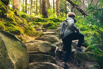 Hiker woman with backpack climbing up a winding path, stones in green woods. Traveling, trekking, recreation, sport, tourism at sunny autumn, spring day. Female traveler on forest trail. Wanderlust.