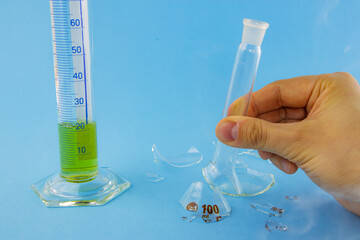 Laboratory experiment. Hand holding broken lab flask glass and measurement glass smoking on blue...