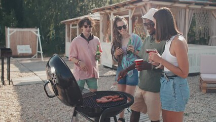 Medium long of African American guy roasting sausages on grill, his young diverse friends talking, drinking and smiling on beach in summer
