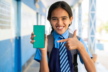 Smiling school girl kid in uniform showing green screen mobile phone by pointing finer while looking camera at corridor - concept of education advertisement and learning application promotion