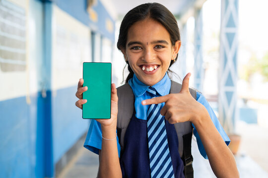 Smiling school girl kid in uniform showing green screen mobile phone by pointing finer while looking camera at corridor - concept of education advertisement and learning application promotion